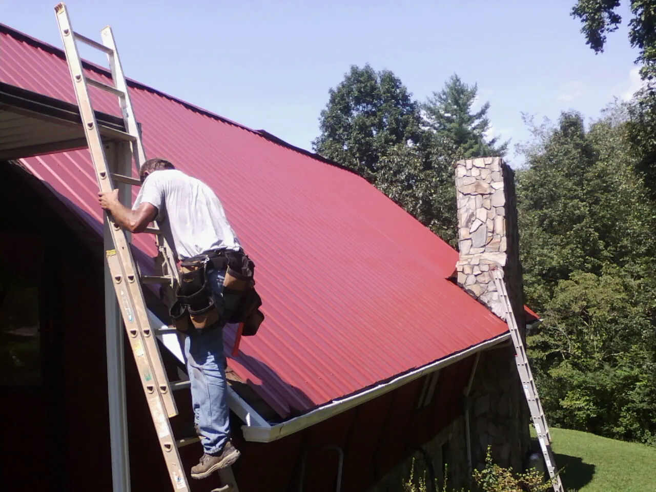 Roofer on a ladder beside a red metal roof and stone chimney — Soffit & Fascia Repair by Roofing Brothers in Welcome